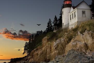 A brick lighthouse on the coast of Maine in Bar Harbor. The foundation is made of stone along a rocky cliff. The restored light glows red against a sunset.