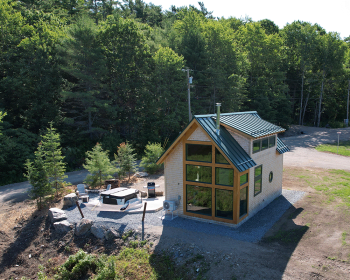 Aerial view of the cabin exterior featuring a large wall of windows, siting amongst Maine's forest, and a hot tub with patio.