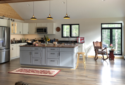 Open, bright newly constructed kitchen with kitchen island built in the center. Large windows showcase the green outdoor living space. White kitchen cabinets have been installed, as well as much more.